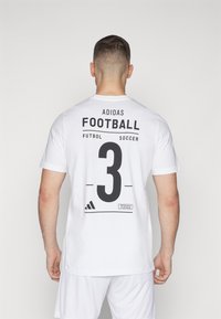 Maglietta bianca a maniche corte con testo nero "ADIDAS FOOTBALL" e numero "3" sul retro, con dettaglio del tag "l'impossibile è nulla".