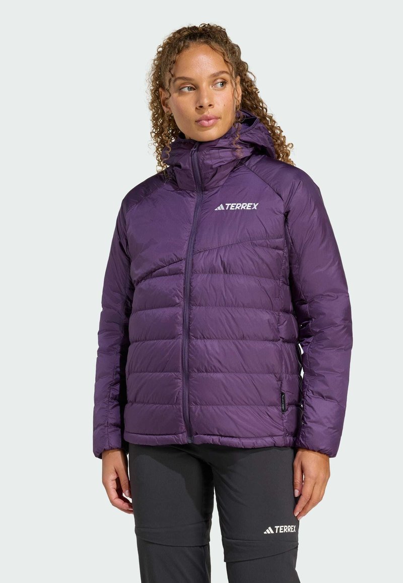 Veste rembourrée violette avec capuche, dotée d'une fermeture éclair et d'une texture gaufrée. Logo sur la poitrine gauche, accompagnée de pantalons sombres.