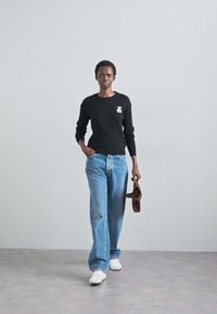Pulover negru cu model canalat și un patch decorativ, blugi largi de culoare albastru deschis și adidași albi. Modelul ține o geantă maro.