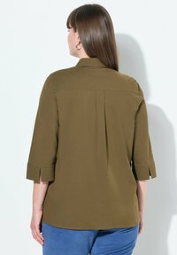 Blusa verde oliva con colletto, maniche a tre quarti, piega sul retro e spacchi laterali. Realizzata in un tessuto liscio con una finitura opaca.