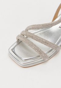 Hopeinen remmikorkokenkä, jossa on neliömäinen kärki, koristeltuna helmikuvioilla remmeissä ja jossa on sileä metallinen pinta.