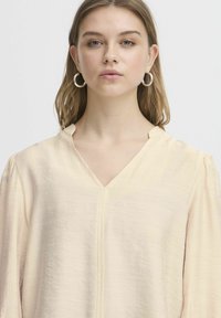 Ljusbeige blus med V-ringning, långa puffärmar och textilstruktur. Har en framseam för ytterligare detalj.