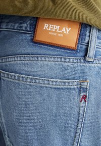 Los jeans de mezclilla azul claro presentan una etiqueta de cuero marrón con "REPLAY SINCE 1981". Unas costuras rojas decoran uno de los bolsillos traseros.