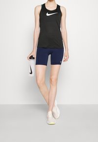 Svart atletisk linne med en vit logotyp, marinblå shorts och ljusa sneakers. Ljus bakgrund, sportvattendosa i handen.