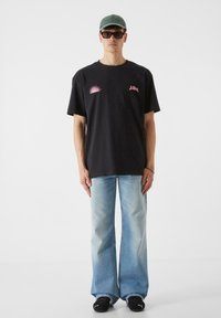 Must musta t-särk, millel on roosa graafiline disain rinnal, koos helesiniste laia säärega teksade ja mustade kingadega. Peas roheline nokamüts ja prillid.