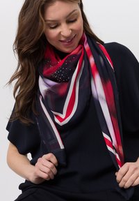 Silk scarf featuring a red, navy, and white geometric pattern with studded accents. Lightweight and soft texture, draped around the neck.