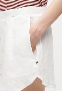 Χέρι μέσα στην τσέπη λινών άσπρων σορτς που φοριούνται με λευκό και κόκκινο ριγέ τοπ, δείχνοντας το ελαστικό ζωνάρι και μια μικρή μαύρη λεπτομέρεια λογότυπου.