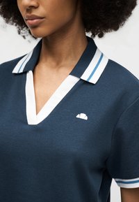 Γυναίκα που φοράει μπλε ναυτικό πόλο πουκάμισο με λευκό και ανοιχτό μπλε ριγέ γιακά και μανσέτες στα μανίκια, με μικρό λευκό λογότυπο στο στήθος.