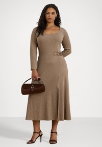 Kvinne iført en langermet beige kjole med sidespenne og slitt, som holder en brun håndveske, og har på seg brune hæler mot en hvit bakgrunn.