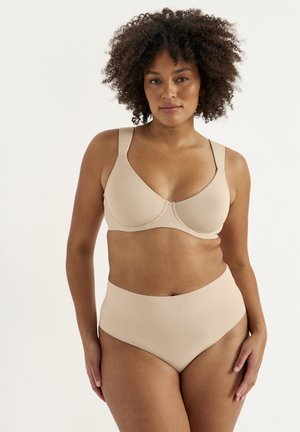 Kvinde med krøllet hår iført beige bh og trusser med høj talje, vendt mod fronten mod en ensfarvet hvid baggrund.