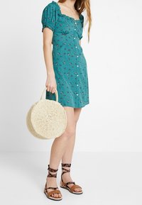 Azurová šaty s květinovým vzorem, nafouknutými rukávy, knoflíky a délkou nad kolena. V kombinaci s kulatou tkanou taškou a sandály na pásky.