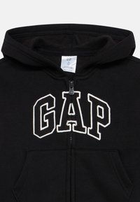 Svart hettejakke for småbarn med hvit omrisset GAP-logo på forsiden, glidelåslukking, kengurulomme og festehette, laget av resirkulert materiale.