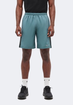 Άνδρας που φοράει τιρκουάζ αθλητικά σορτς με ελαστική μέση, μαύρα αθλητικά παπούτσια, λευκές κάλτσες Nike, στέκεται μπροστά σε απλό ανοιχτό γκρι φόντο.