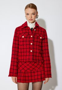 Giacca di tweed rossa con un motivo a scacchi neri, dotata di colletto, bottoni di perla e un design corto. Abbinata a una gonna coordinata.