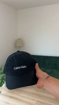 Svart baseballcap laget av stoff med buet brem, med hvit brodert "Calvin Klein"-logo på forsiden. Enkel design.