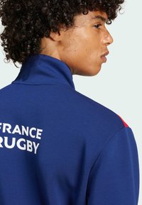 Blaue Rugbyjacke mit hohem Kragen, versehen mit "FRANCE RUGBY" in weißem Text. Rote Akzente an den Schultern. Glatte Textur des Stoffes.