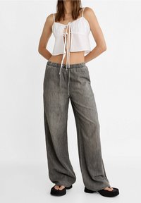Bílý cropped top s tenkými ramínky a detaily vázání vpředu, spárovaný s volnými šedými kalhotami s elastickým pasem a rovnými nohavicemi.