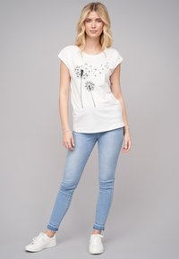 T-shirt blanche à manches courtes avec un graphique de pissenlit noir, accompagnée d'un jean skinny bleu clair et de baskets blanches. Fond uni.