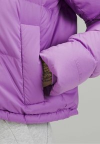 Doudoune violette avec un design matelassé, dotée d'une poche latérale, de poignets élastiques et d'un logo discret sur la manche. Texture lisse.