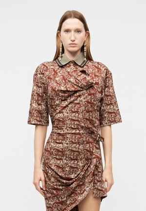 Sieviete tērpusies rūsganā krāsā kleitā ar leoparda rakstu un zaļu apkakli, ar auduma drapējuma detaļām un garām pērļu auskariem.