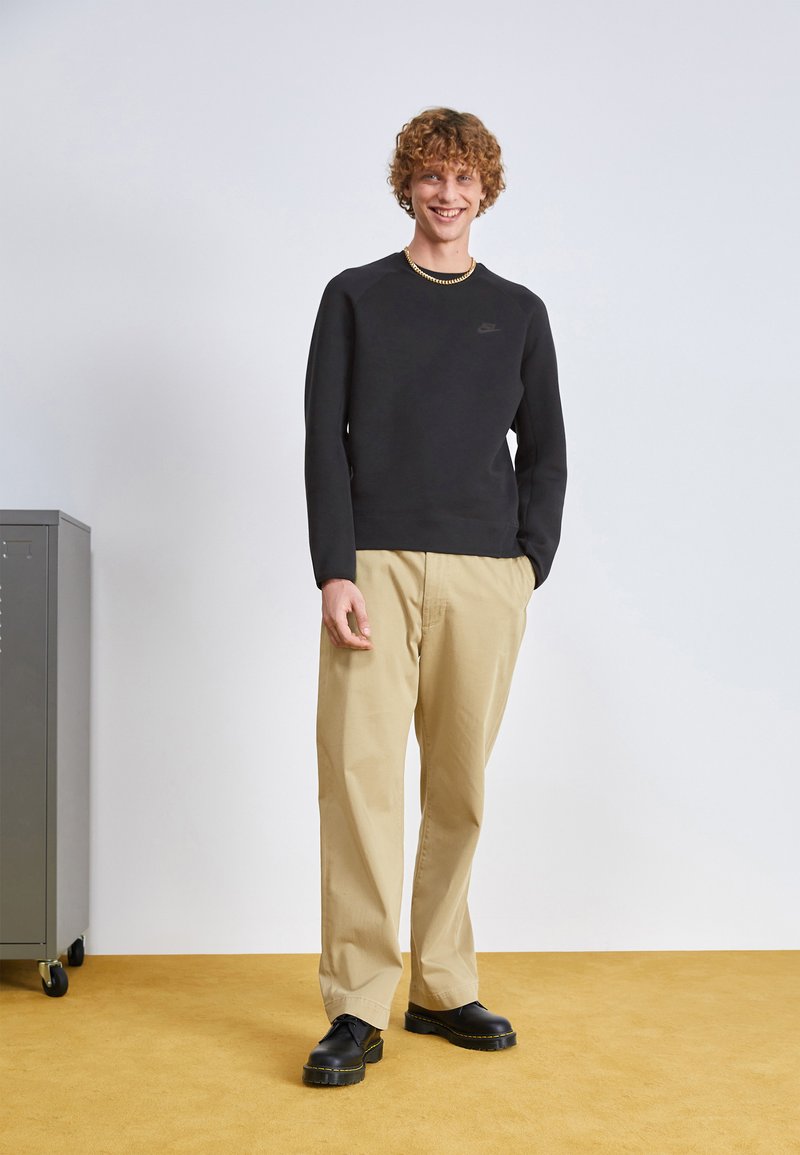 Jeune homme souriant, portant un sweat-shirt noir, un pantalon beige, des chaussures noires, se tenant sur un sol jaune à côté d'un cabinet gris sur roues.