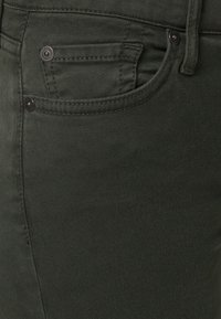 Jeans de mezclilla verde oscuro con un bolsillo frontal con detalles de costura y botones de metal negro, textura suave y un diseño ajustado.