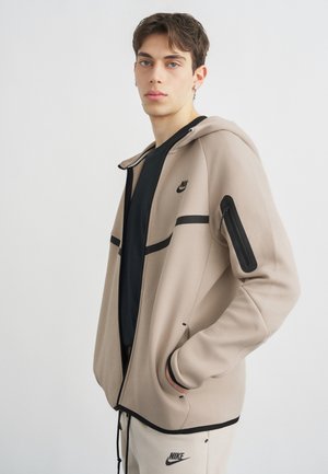 Giovane con capelli corti che indossa una felpa Nike beige con zip e pantaloni della tuta abbinati, posando con le mani nelle tasche su sfondo bianco.