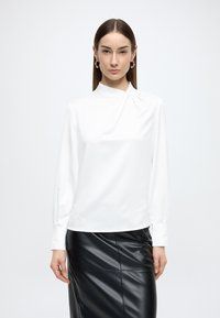 Bluză albă cu mâneci lungi și guler înalt, având un detaliu drapat la guler și manșete strânse, purtată băgată într-o fustă neagră din piele.