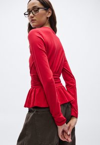 Rødt langermet topp med innsvingt midje og peplumkant, glatt tekstur og tettsittende silhuett, brukt sammen med mørke bukser.