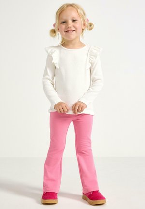 Långärmad vit topp med volanger vid axlarna, kombinerad med rosa utsvängda byxor och rosa fluffiga skor. Enkelt mönster och mjuka material.