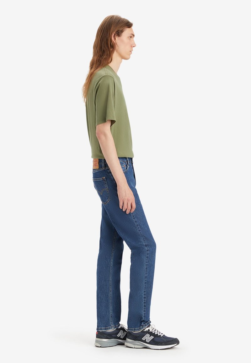 T-shirt à manches courtes vert olive, jean en denim bleu et baskets noires. Design simple, coupe droite, avec des coutures visibles et un détail d'étiquette.