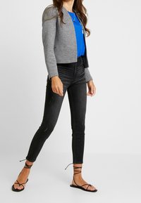 Veste grise courte portée sur un haut bleu, associée à un jean skinny noir et des sandales noires à lanières. Texture mate avec des détails minimaux.