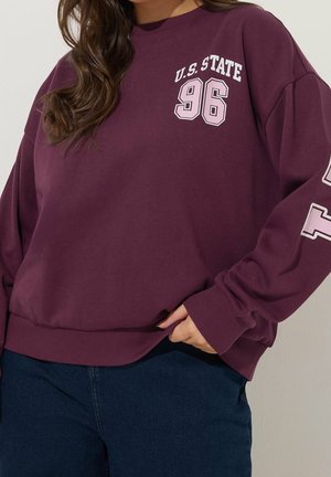 Woman wearing a maroon sweatshirt with "U.S. STATE 96" text on chest and sleeve, paired with dark blue jeans, hand resting on hip.