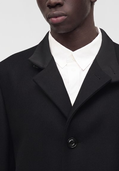 Gros plan d'une personne portant un manteau noir avec des revers en satin sur une chemise blanche à col, montrant la partie inférieure du visage et le cou.