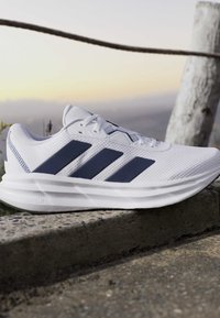 Chaussure de sport blanche avec des rayures et lacets bleu marine reposant sur une surface en pierre avec un arrière-plan extérieur flou au lever ou au coucher du soleil.