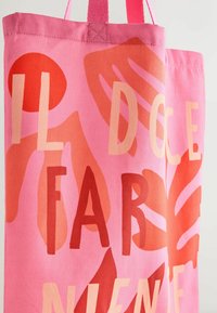 Pink fabric tote bag with red and orange abstract leaf patterns and partially visible large, bold letters in beige and red.