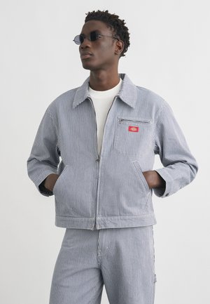 Hombre con chaqueta con cremallera de rayas finas azules y blancas y pantalones a juego, con gafas de sol redondas negras, manos en los bolsillos, mirando hacia la izquierda.