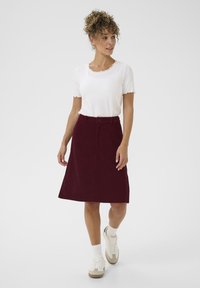 Jupe en velours côtelé bourgogne en forme A avec poches avant et passants de ceinture, associée à un haut blanc à manches courtes en côtelé et des baskets blanches.