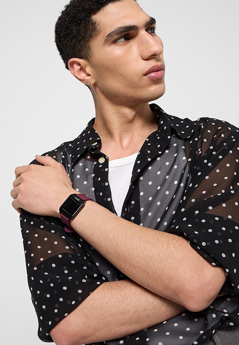 Transparente schwarze Bluse mit weißen Pünktchen, die über einem weißen T-Shirt getragen wird, und einer burgunderroten Smartwatch am Handgelenk.