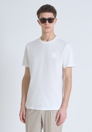 Weißes Baumwoll-T-Shirt mit kurzen Ärmeln, Rundhalsausschnitt und strukturiertem Bärenlogo auf der linken Brust, kombiniert mit hellbeigen Hosen.