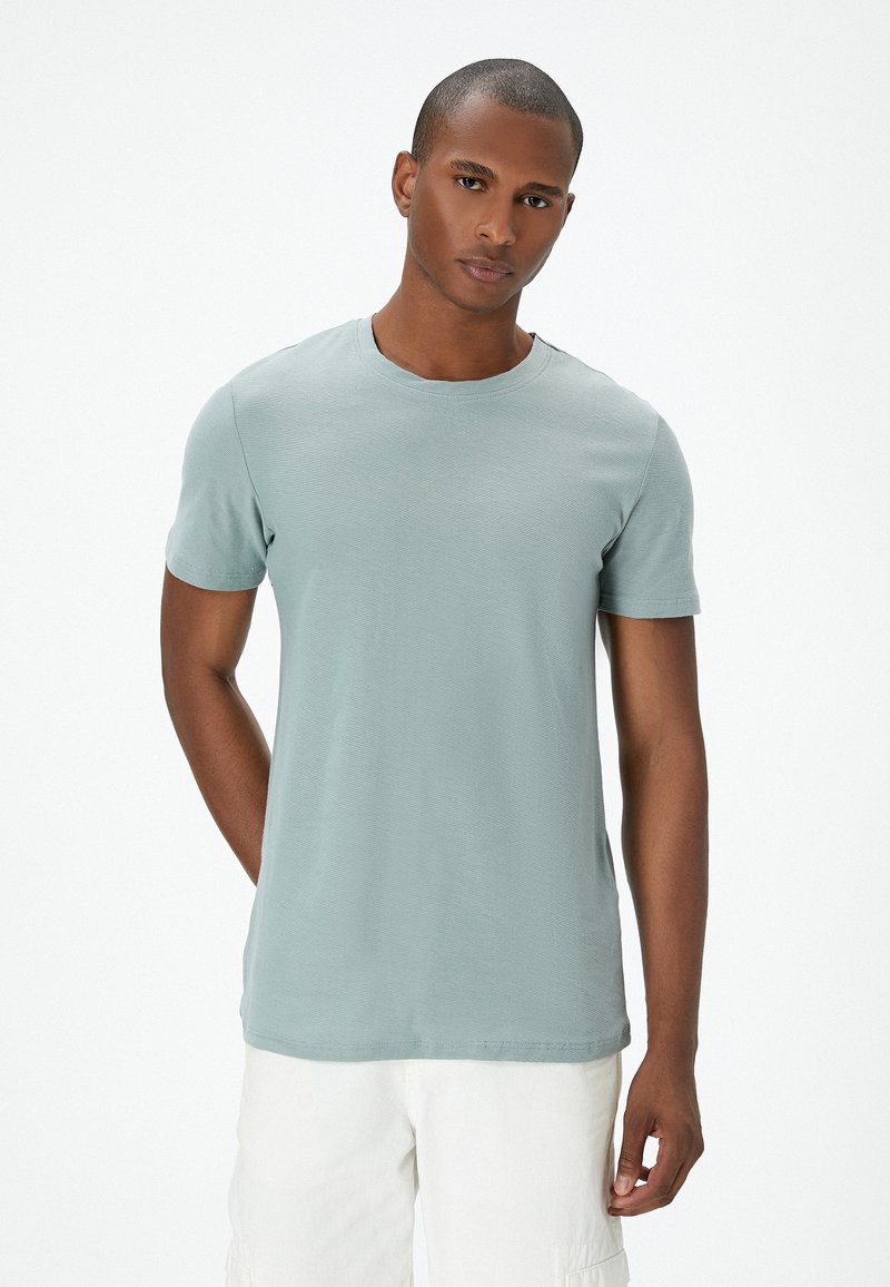 Lyseblåt kortærmet t-shirt med rund hals, lavet af et struktureret stof. Modellen står med hænderne i lommerne foran en hvid baggrund.