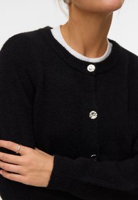 Femme portant un cardigan en maille noire avec des boutons ronds argentés brillants sur un haut blanc à col rond, bras croisés montrant une bague en argent.