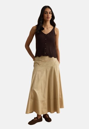 Frau mit langen dunklen Haaren trägt ein ärmelloses braunes Oberteil mit Knöpfen, einen beigen knöchellangen Rock, ein goldenes Armband und dunkelbraune Schuhe und steht selbstbewusst da.