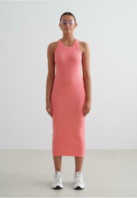 Rochie fără mâneci, roz, cu model în relief, cu o siluetă ajustată, până la genunchi. Pură cu adidași albi, pe un fundal simplu, de culoare deschisă.