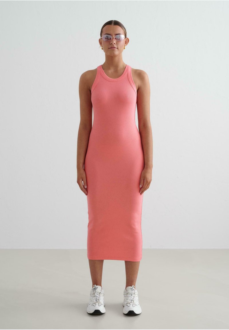 Rochie fără mâneci, roz, cu model în relief, cu o siluetă ajustată, până la genunchi. Pură cu adidași albi, pe un fundal simplu, de culoare deschisă.