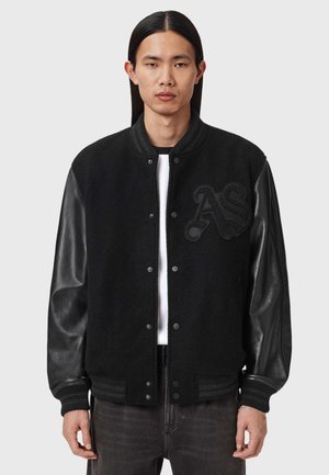 Mężczyzna z długimi czarnymi włosami ma na sobie czarną kurtkę varsity z leatherowymi rękawami i czarne tłoczone logo "AS" na piersi, na białej koszuli.