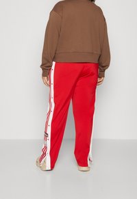 Person som bär röda träningsbyxor med vita sidostripes och knappar, brun sweatshirt och beige skor, står på ett enkelt ljust golv.