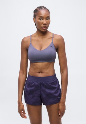 Femme aux cheveux tressés portant un soutien-gorge de sport violet et un short de sport violet foncé, debout devant un fond uni.