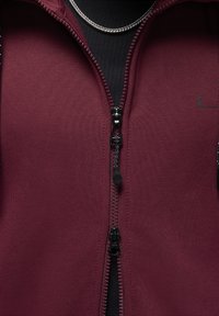 Gros plan d'une veste bordeaux zippée avec tirant de fermeture noir et collier chaîne argenté sur chemise noire.