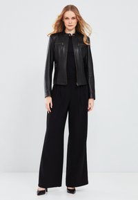 Black leather jacket with zippered pockets, round collar, and fitted design, worn over a black jumpsuit and paired with black heels.